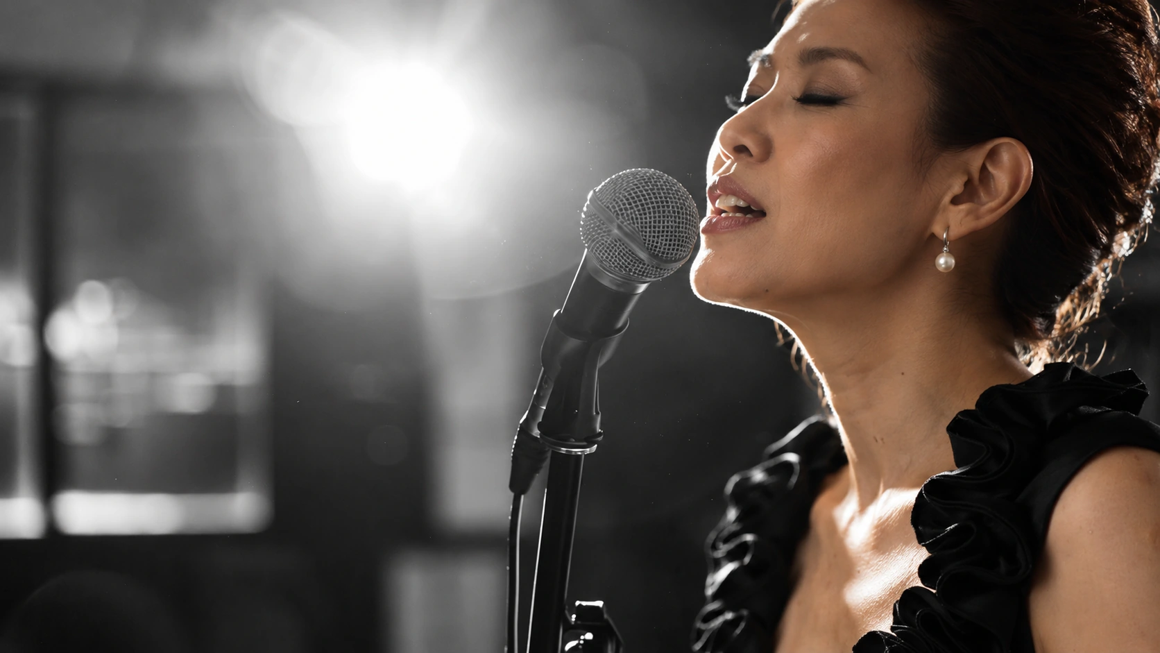 A woman singing into a microphone in natural colour, against a monochrome studio with dramatic stage lighting.
